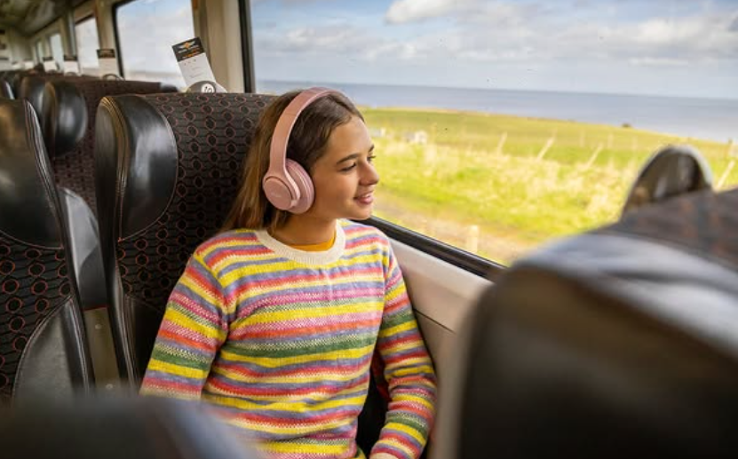 girl sat on a train with headphones on smiling whilst staring out of the window